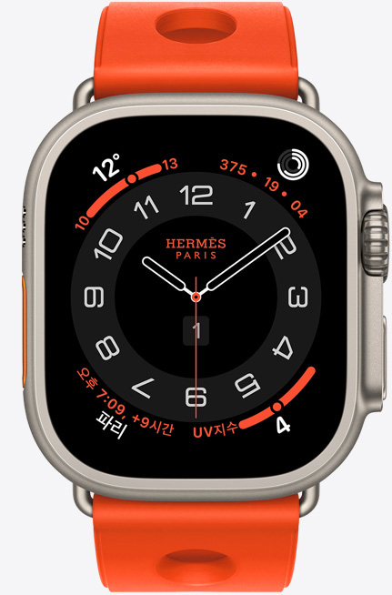 Apple Watch Hermès Ultra 3 Circulaire 아날로그 시계 페이스에 (시, 분, 초침, 컴플리케이션: 날씨, 활동 링, 파리 시간 및 자외선 지수), 내추럴 색상 티타늄 케이스, 오른쪽 옆면 외관, Digital Crown, 돌출된 측면 버튼, 오랑쥬 네옹(밝은 오렌지) 색상의 Scub’H Diving 밴드(중앙에 구멍이 뚫린 성형 고무 소재)와 연결되어 있다.