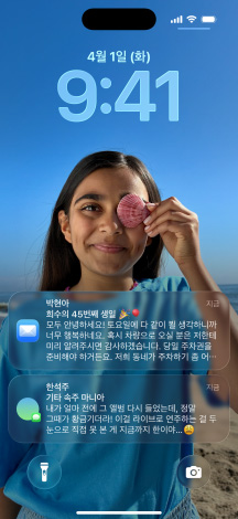 iPhone, 다섯 가지 모델, 각 모델은 옆에 있는 모델과 약간 겹치며, 맨 왼쪽 화면에는 배경이 있는 대화가 표시된 메시지 앱, 왼쪽에서 두 번째 화면에는 다채로운 앱 아이콘이 있는 홈 화면, 가운데 모델에는 사진이 있는 잠금 화면과 Liquid Glass로 표시된 시간 숫자와 화면 하단에 표시된 두 개의 알림, 오른쪽에서 두 번째 모델에는 투명한 Liquid Glass 앱 아이콘, 맨 오른쪽 모델에는 Liquid Glass로 표시된 음악 플레이어가 있는 모습
