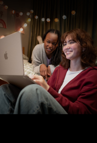 Two students view content on a silver MacBook in their dorm room, with one sitting on the bed as the other sits on the floor holding the device