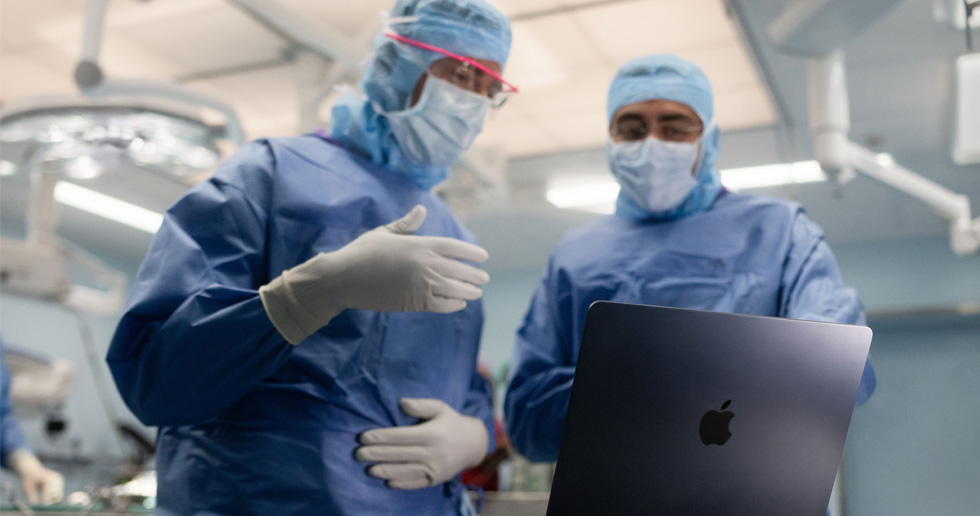 Deux chirurgiens dans une salle d’opération regardent un MacBook, l’un fait un geste de la main tandis que l’autre observe