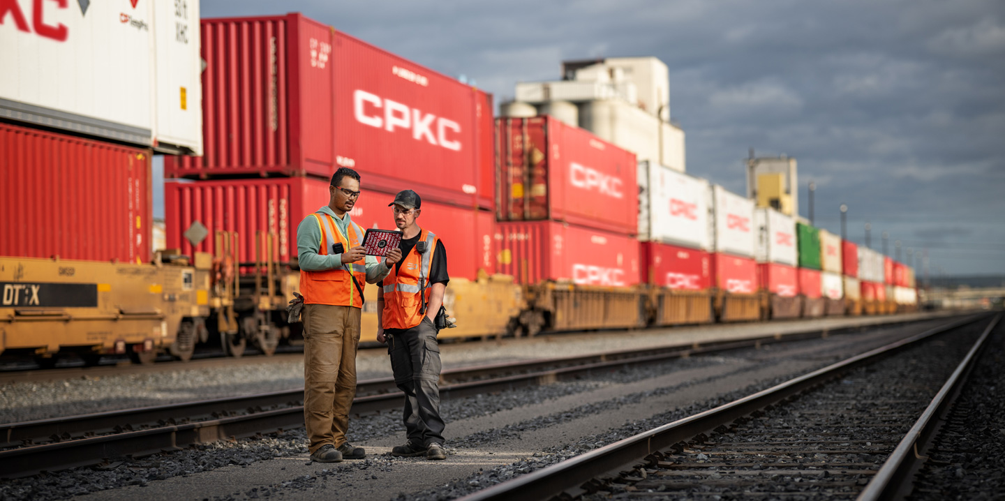 Deux employés des chemins de fer se trouvent dans une gare de triage et consultent un iPad à côté d’un train à l’arrêt transportant des conteneurs d’expédition