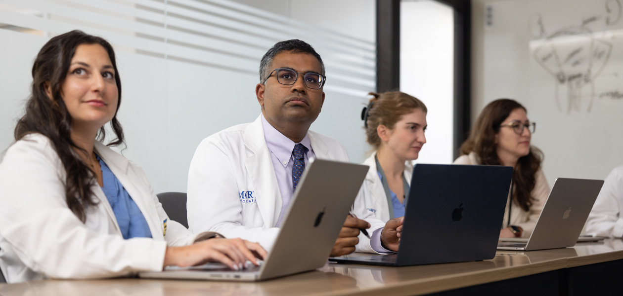 Four colleagues in doctors' coats sit in row, each working on a MacBook.